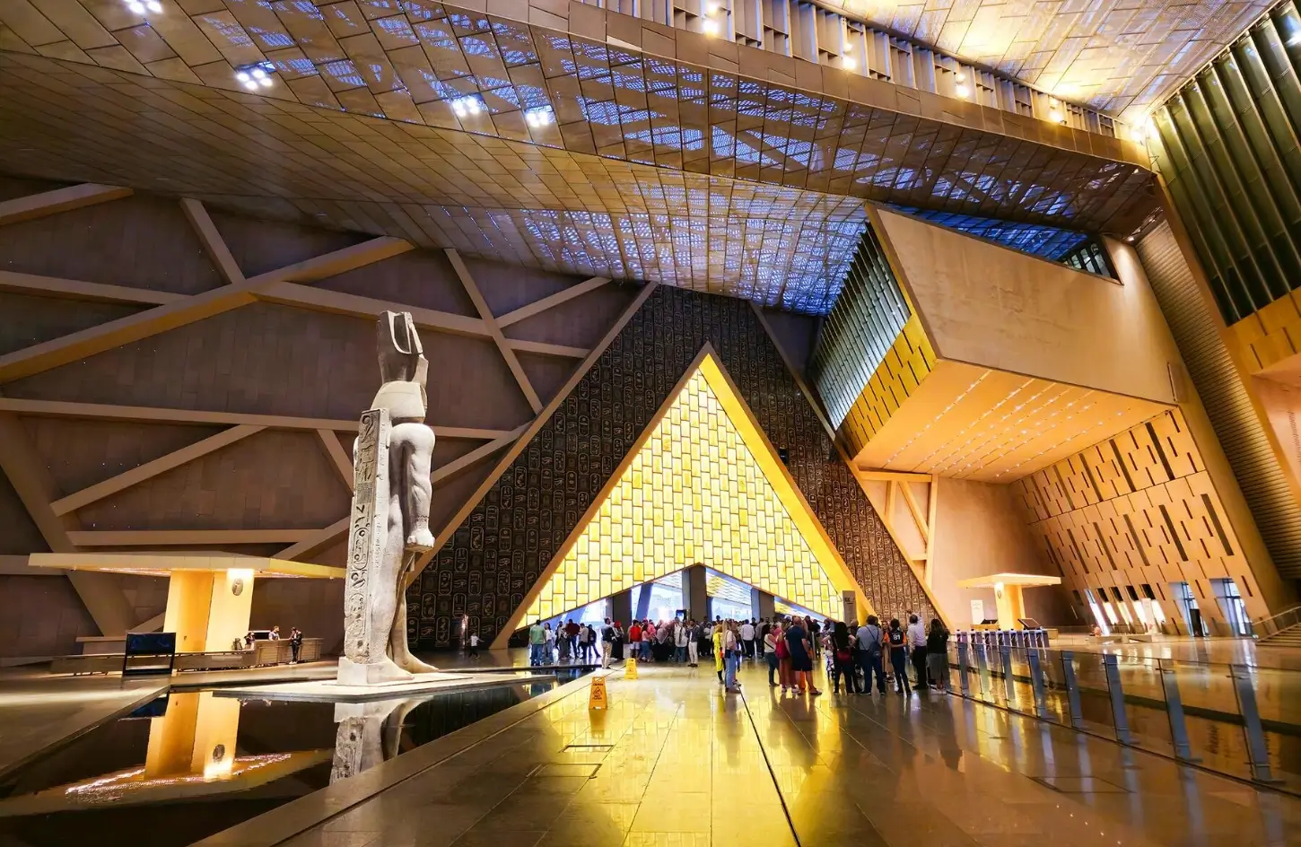 Visitors gathering near a large ancient statue and illuminated triangular entrance portal inside the Grand Egyptian Museum.